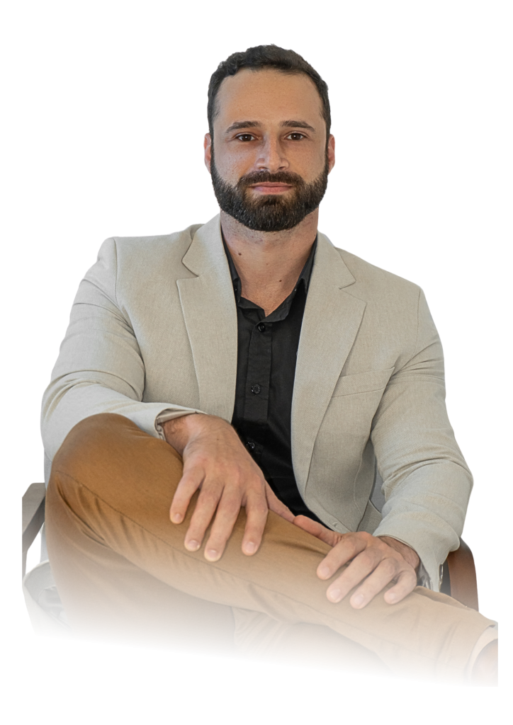 Homem sentado em uma cadeira, vestindo blazer bege, camisa preta e calça marrom, com expressão séria e pernas cruzadas.
