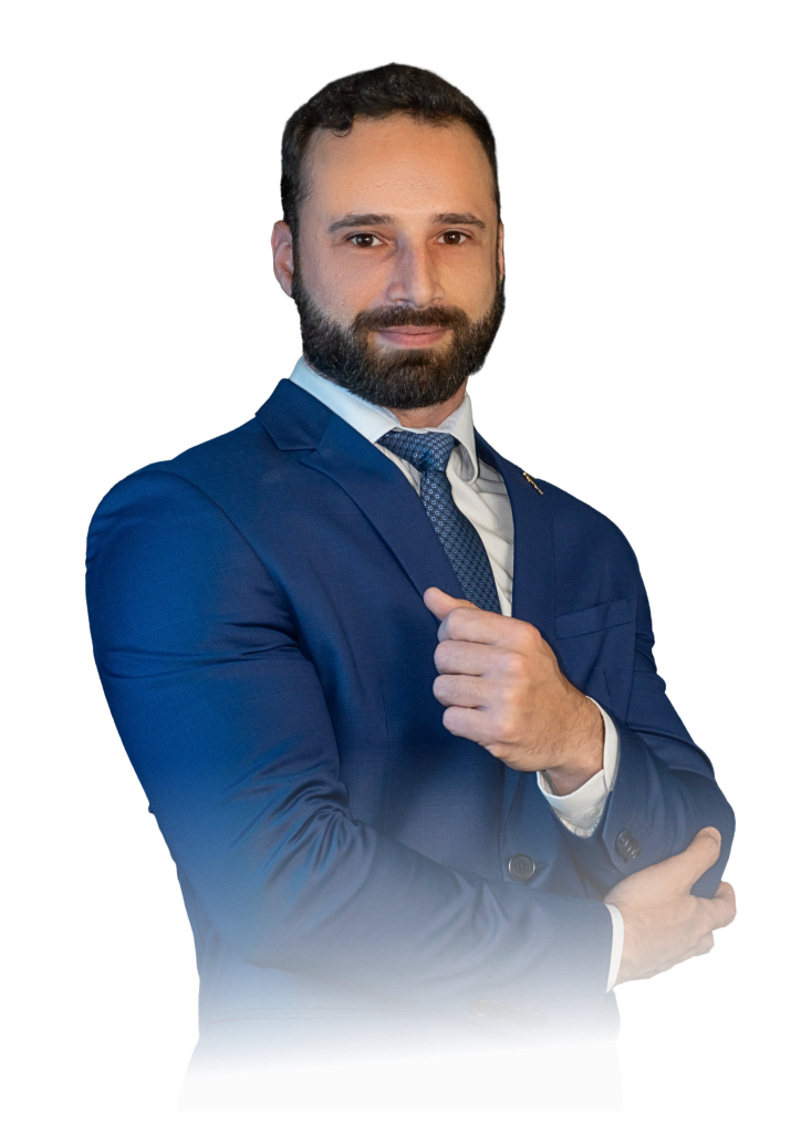 Homem em pé, vestindo terno azul, camisa branca e gravata azul, olhando para a câmera com expressão confiante e ajustando a manga do paletó.